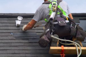 Licensed roofer replacing shingles safely using harness and elevated platform