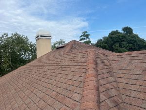 Wide view of an aging reddish-brown shingle roof with a stucco chimney and tree coverage in Sawgrass, FL.